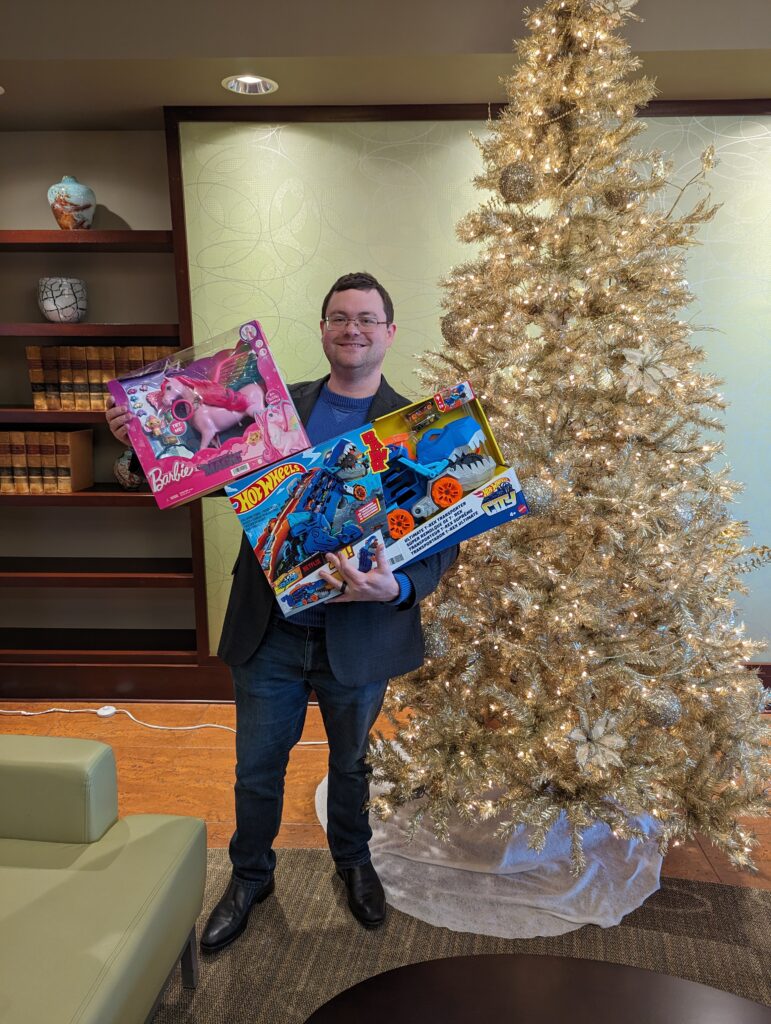 Man holding gifts in front of a Christmas tree for donation to Toys for Tots.